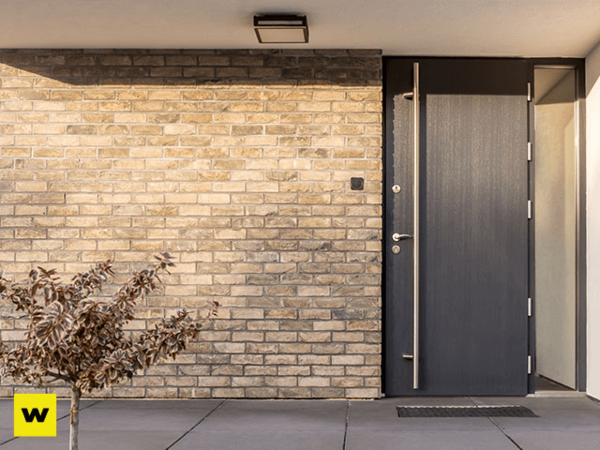 Moderne Haustür aus Aluminium neben heller Klinkerfassade und kleinem Baum im Eingangsbereich.